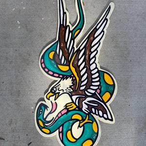 May include: A green and yellow snake wrapped around a brown and white eagle with its wings spread. The eagle's beak is open and the snake's tail is curled around the eagle's neck. The image is painted on a wooden surface.