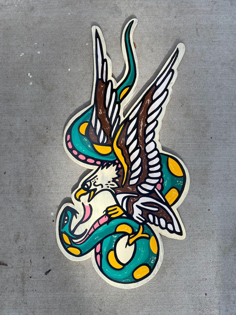 May include: A green and yellow snake wrapped around a brown and white eagle with its wings spread. The eagle's beak is open and the snake's tail is curled around the eagle's neck. The image is painted on a wooden surface.