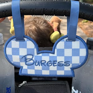 May include: A blue and white checkered Mickey Mouse ear-shaped stroller tag with the name "Burgess" embroidered in blue. The tag is attached to a stroller handle with blue straps. The background is blurred.