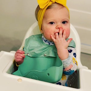 May include: A baby in a high chair wearing a yellow bow headband, a light green bib, and a patterned long-sleeved shirt. The baby is holding a small stick and has food on their face.