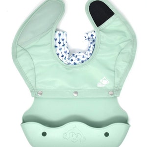 May include: A mint green baby bib with a built-in food catcher. The bib has a patterned interior with blue and white arrows. The bib has a monkey logo and snaps for closure. The bib is made of a soft, waterproof material.