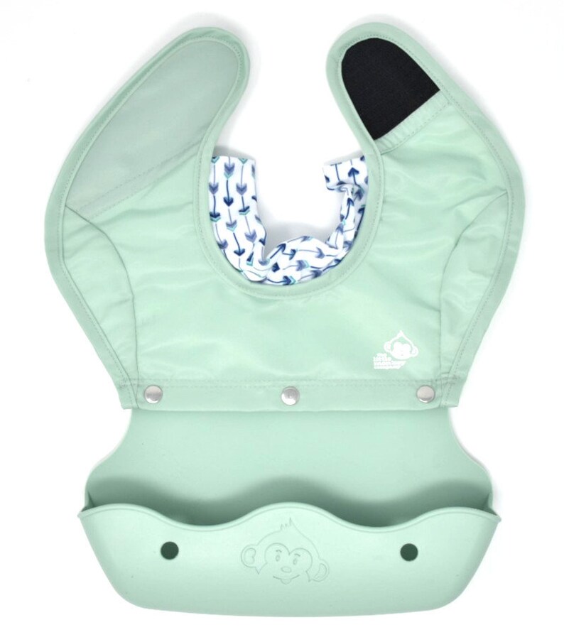 May include: A mint green baby bib with a built-in food catcher. The bib has a patterned interior with blue and white arrows. The bib has a monkey logo and snaps for closure. The bib is made of a soft, waterproof material.