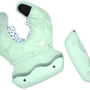 May include: A mint green baby bib with a silicone food catcher and a detachable sleeve. The bib has a monkey logo and a patterned interior with blue arrows on a white background. The sleeve is also mint green.