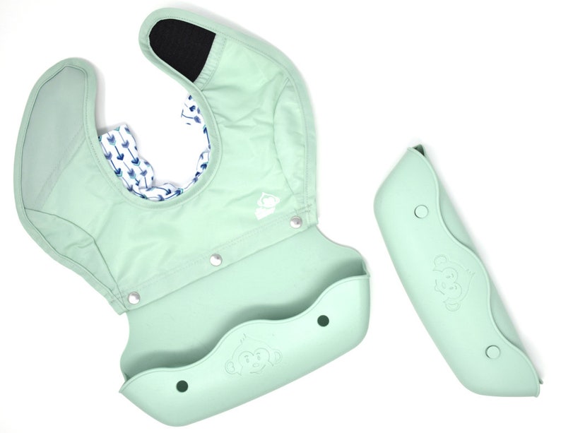 May include: A mint green baby bib with a silicone food catcher and a detachable sleeve. The bib has a monkey logo and a patterned interior with blue arrows on a white background. The sleeve is also mint green.