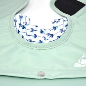 May include: A mint green baby bib with a white and blue arrow pattern on the inside collar. The bib has a snap closure and a velcro closure on the shoulder straps. The Little Monkey Company logo is visible.