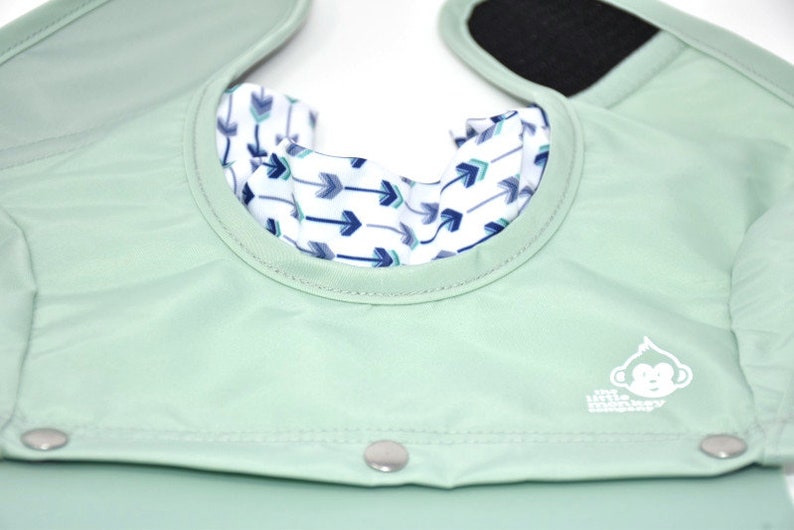 May include: A mint green baby bib with a white and blue arrow pattern on the inside collar. The bib has a snap closure and a velcro closure on the shoulder straps. The Little Monkey Company logo is visible.