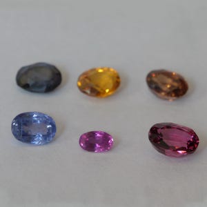 Peut inclure: Six pierres précieuses de forme ovale de différentes couleurs, notamment bleu, jaune, rose et marron. Les pierres précieuses sont disposées en rangée sur une surface blanche.