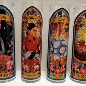 May include: Four glass candle holders with colorful stained glass designs. Each candle holder features a different image and text. The first candle holder has a black and white image of a robot with the text "I Am Still Alive". The second candle holder has a colorful image of a woman with the text "Unlikeable, What Not Be More". The third candle holder has a colorful image of a cube with the text "Your Best Friend". The fourth candle holder has a colorful image of a cake with the text "The Cake Is A Lie".