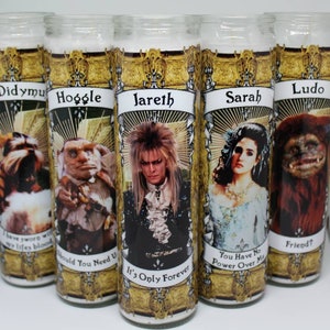 May include: Five white glass candle jars with gold trim and colorful images of characters from the movie Labyrinth. The candles feature the characters Sir Didymus, Hoggle, Jareth, Sarah, and Ludo. Each candle has a quote from the movie printed below the image.
