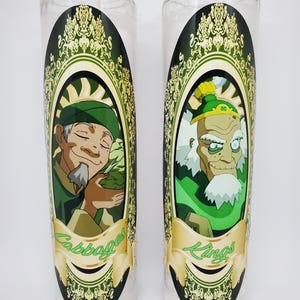 May include: Two white glass candle holders with green and gold designs. The left candle holder features a cartoon character with a green hat and the text "Cabbages". The right candle holder features a cartoon character with a green hat and the text "Kings".