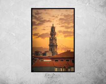 Pode incluir: Uma alta torre de igreja adornada com um relógio de face se destaca contra um fundo de céu de pôr do sol laranja e rosa vibrante. A torre é cercada por telhados de telhas vermelhas de edifícios em uma cidade.