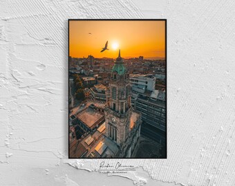 Puede incluir: Fotografía enmarcada de un paisaje urbano al atardecer. La imagen presenta un edificio alto y ornamentado con una cúpula verde, sobre un cielo naranja vibrante. Un pájaro vuela cerca del sol, y la ciudad se extiende debajo.