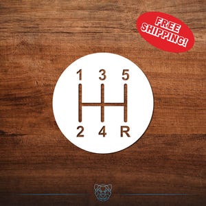 May include: White circular graphic of a car's manual transmission gear shift pattern, with numbers 1 through 5 and R for reverse. The graphic is on a dark wood grain background. A red oval sticker with the text "FREE SHIPPING!" is in the upper right corner.