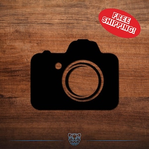 May include: A black silhouette of a camera on a wooden background. The camera shape is simple, with a lens and a button. A red oval sticker with the words "FREE SHIPPING!" is in the upper right corner.