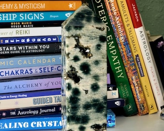 May include: A green and white crystal point with a unique marbled pattern. The crystal is standing upright on a bookshelf with several books about astrology and mysticism.