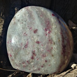May include: A large, smooth, white and gray polished stone with red and pink inclusions. The stone has a rounded shape and is sitting on a piece of bark.