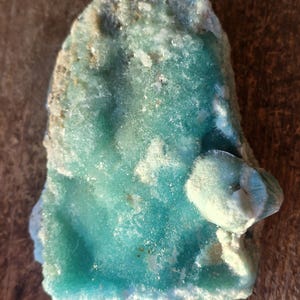 Puede incluir: Esp&eacute;cimen mineral natural de forma irregular, con un vibrante tono turquesa. La superficie tiene una apariencia texturizada y cristalina con &aacute;reas blancas y marr&oacute;n claro. El mineral se encuentra sobre una superficie de madera.