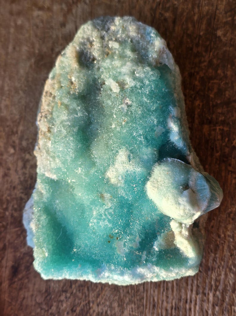Puede incluir: Esp&eacute;cimen mineral natural de forma irregular, con un vibrante tono turquesa. La superficie tiene una apariencia texturizada y cristalina con &aacute;reas blancas y marr&oacute;n claro. El mineral se encuentra sobre una superficie de madera.