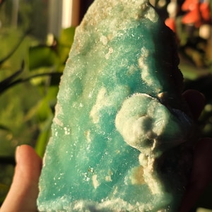 Puede incluir: Una mano sostiene un esp&eacute;cimen mineral grande con bordes &aacute;speros. La piedra es principalmente de color turquesa vibrante con parches blanquecinos y beige. La superficie tiene una textura cristalina y la forma general es irregular.