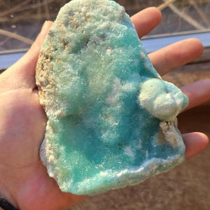 Puede incluir: Un trozo de mineral natural en bruto con un llamativo tono turquesa. La piedra tiene una superficie rugosa y texturizada con &aacute;reas de formaciones cristalinas m&aacute;s claras, casi blancas. El mineral se sostiene en una mano.