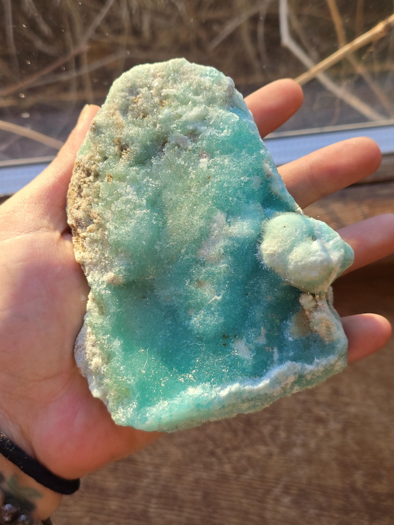 Puede incluir: Un trozo de mineral natural en bruto con un llamativo tono turquesa. La piedra tiene una superficie rugosa y texturizada con &aacute;reas de formaciones cristalinas m&aacute;s claras, casi blancas. El mineral se sostiene en una mano.