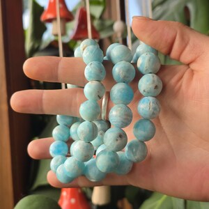 May include: A collection of light blue beaded bracelets. The beads are round and have a marbled appearance. The bracelets are held in a hand, with a silver ring visible. The background includes decorative mushroom ornaments and greenery.