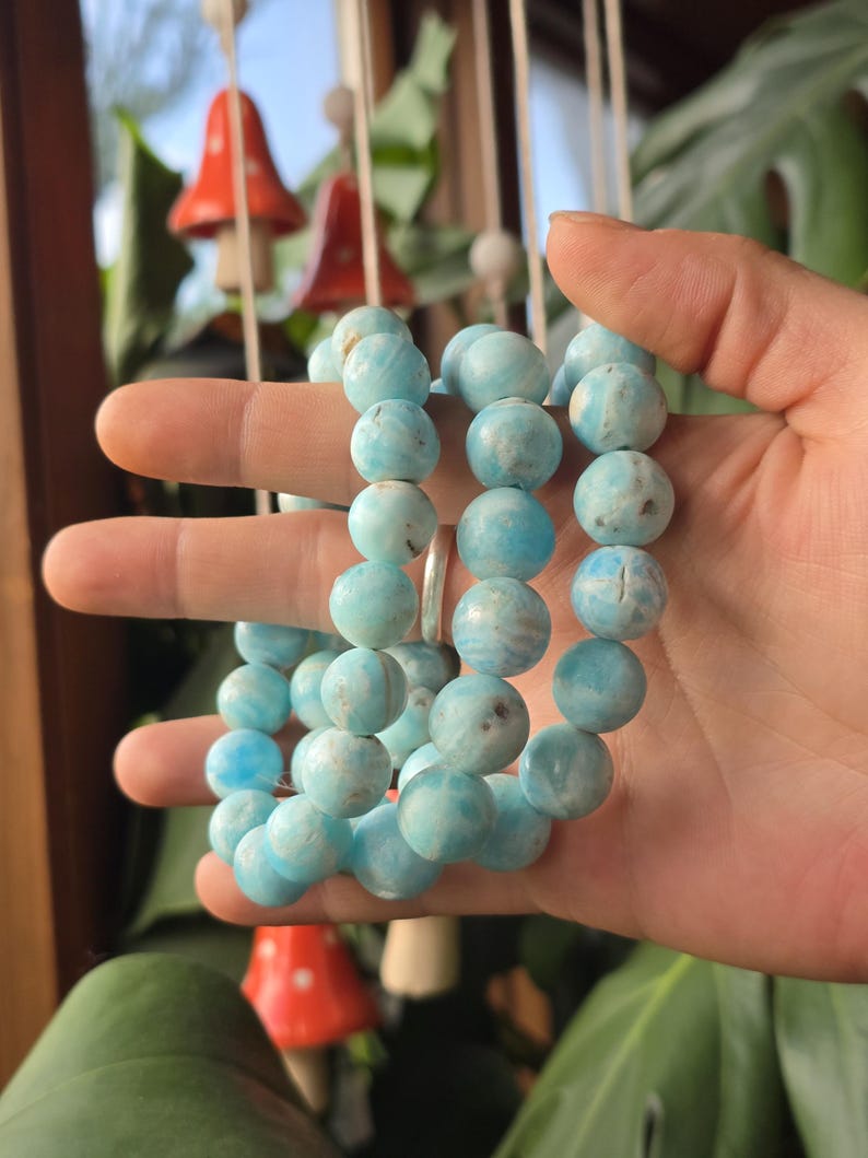 May include: A collection of light blue beaded bracelets. The beads are round and have a marbled appearance. The bracelets are held in a hand, with a silver ring visible. The background includes decorative mushroom ornaments and greenery.