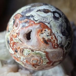Può includere: Una pietra sferica lucidata con aspetto di geode. La pietra presenta un mix di colori, tra cui bianco, arancione, verde e grigio, con una texture superficiale screpolata. Una cavità è visibile al centro. Questo oggetto decorativo è un pezzo unico per l'arredamento della casa.