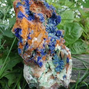 May include: A cluster of blue azurite, green malachite, and clear quartz crystals on a rough, orange-brown rock. The crystals are clustered together in a natural formation.