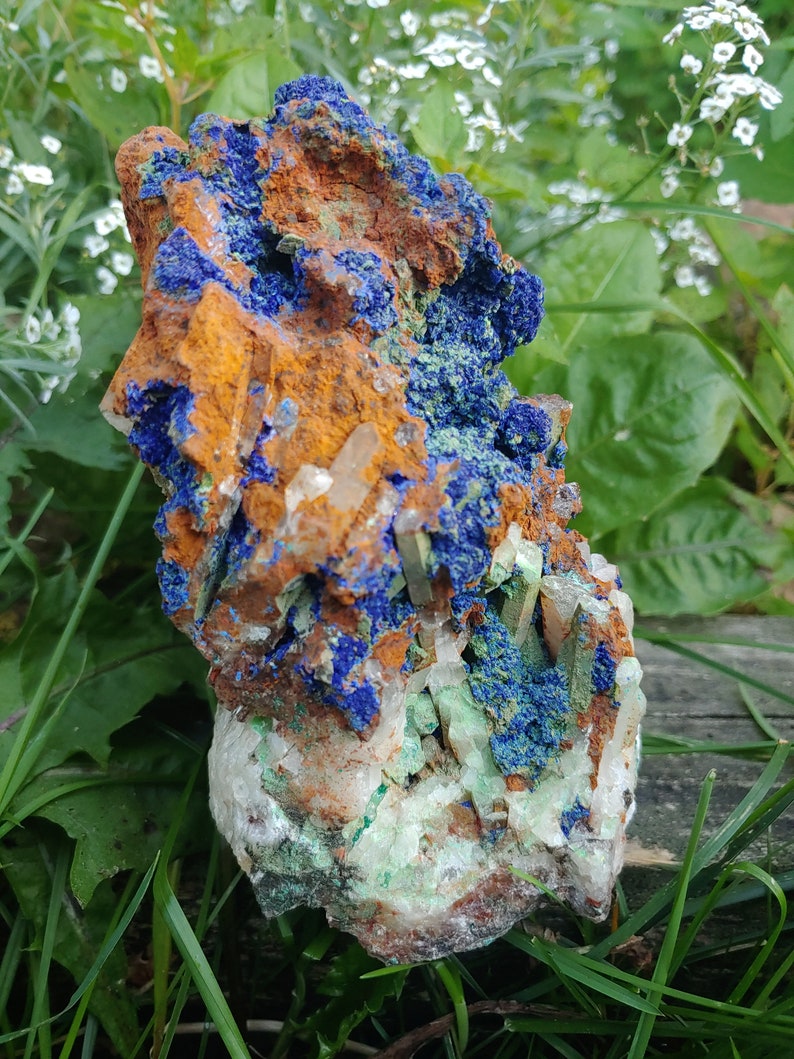 May include: A cluster of blue azurite, green malachite, and clear quartz crystals on a rough, orange-brown rock. The crystals are clustered together in a natural formation.