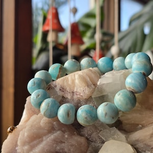 May include: A turquoise beaded bracelet rests on a bed of crystals and minerals. The bracelet features large, round beads with a marbled appearance. The background includes blurred decorative elements and greenery, creating a natural aesthetic.