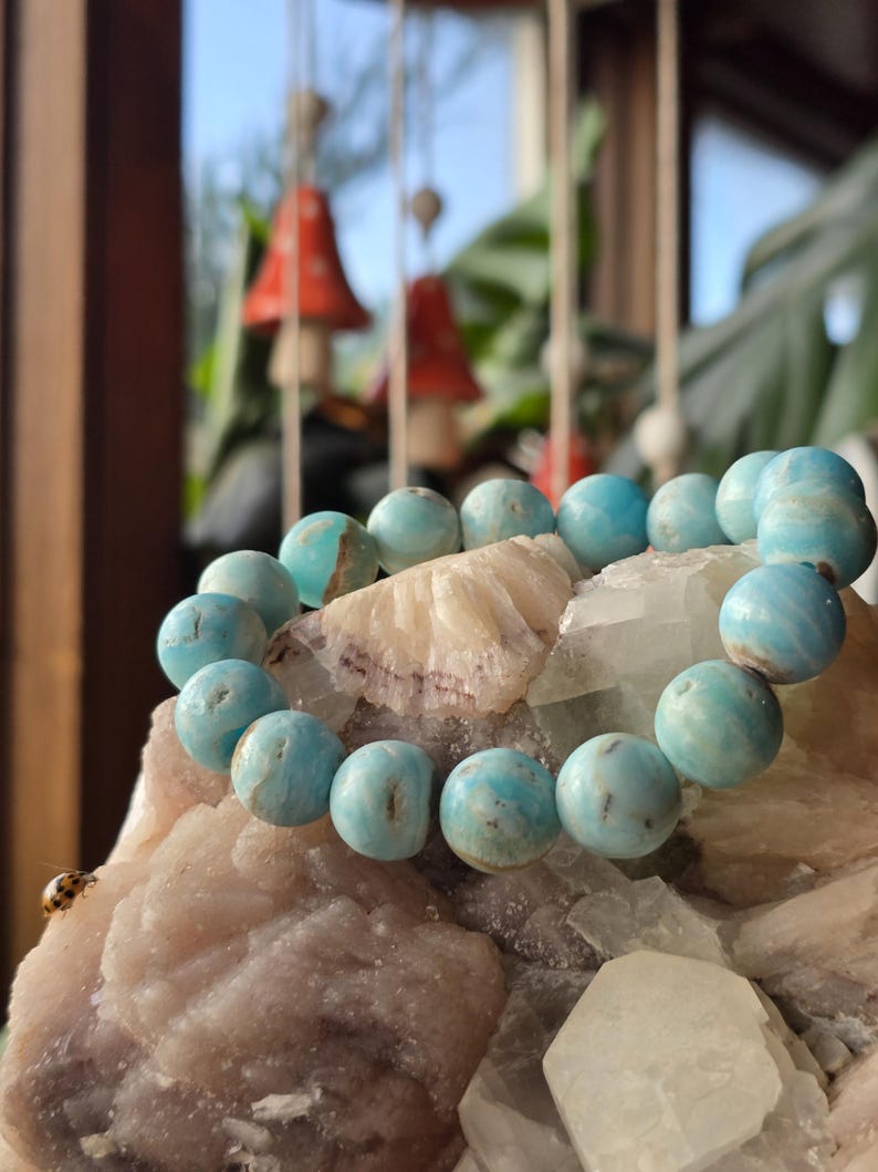 May include: A turquoise beaded bracelet rests on a bed of crystals and minerals. The bracelet features large, round beads with a marbled appearance. The background includes blurred decorative elements and greenery, creating a natural aesthetic.