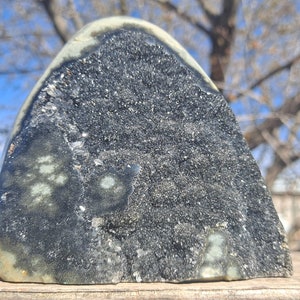 May include: A large, dark grey geode with a natural, rough exterior and a sparkling, crystalline interior. The geode is partially open, revealing the intricate crystal formations inside. The geode is sitting on a wooden surface.