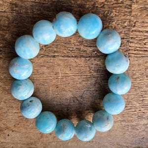 May include: A round bracelet made of light blue, spherical beads. The beads have a marbled appearance with darker blue and white swirls. The bracelet is displayed on a wooden surface.