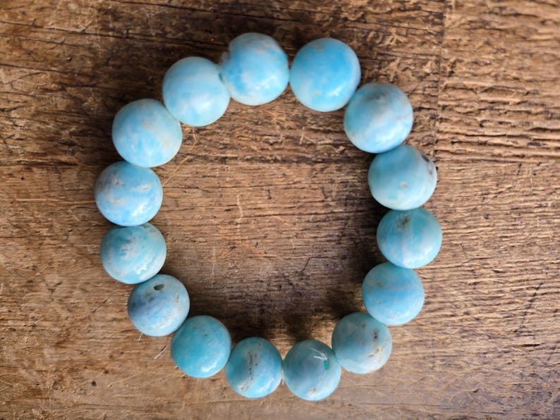 May include: A round bracelet made of light blue, spherical beads. The beads have a marbled appearance with darker blue and white swirls. The bracelet is displayed on a wooden surface.