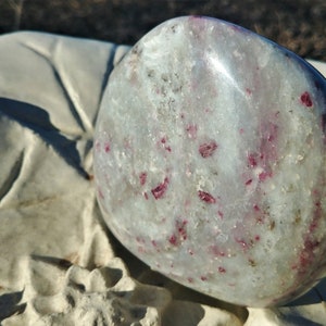 May include: A smooth, white, polished stone with red ruby inclusions. The stone is oval shaped and has a glossy finish.