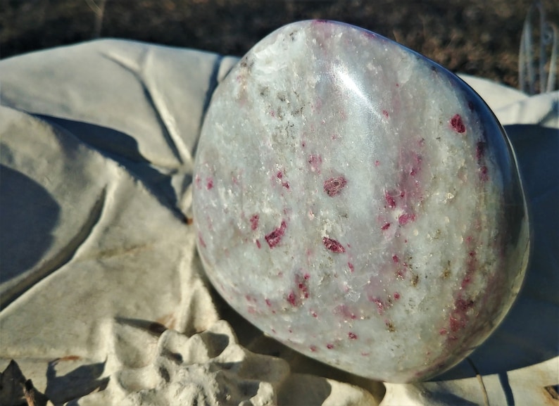 May include: A smooth, white, polished stone with red ruby inclusions. The stone is oval shaped and has a glossy finish.