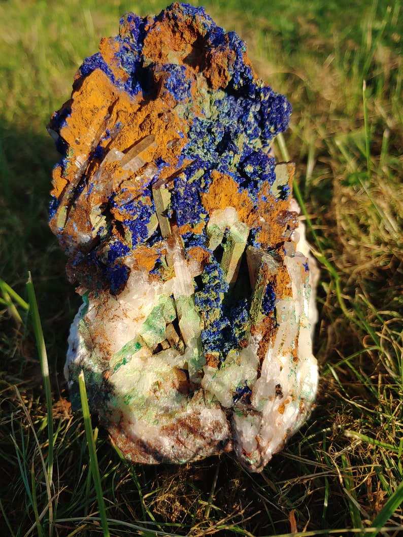 May include: A cluster of blue azurite, green malachite, and clear quartz crystals. The crystals are embedded in a matrix of brown iron oxide.