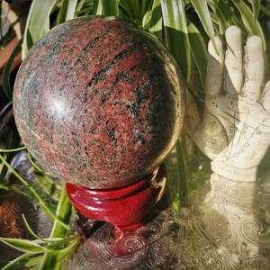 May include: A large, polished, red and green sphere on a red wooden stand. The sphere is made of a type of stone with a swirling pattern.