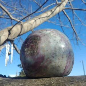 May include: A smooth, round, polished stone with swirls of purple, gray, and white. The stone is resting on a branch with a blue sky and bare tree branches in the background.