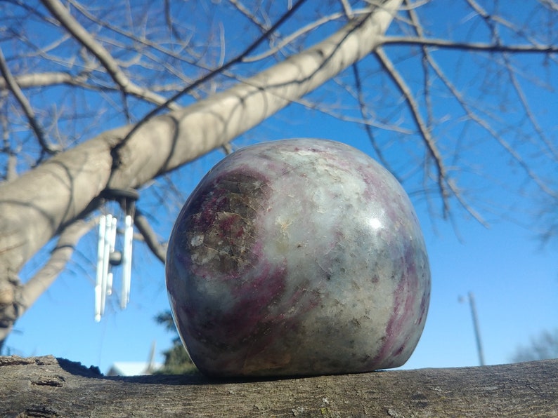 May include: A smooth, round, polished stone with swirls of purple, gray, and white. The stone is resting on a branch with a blue sky and bare tree branches in the background.