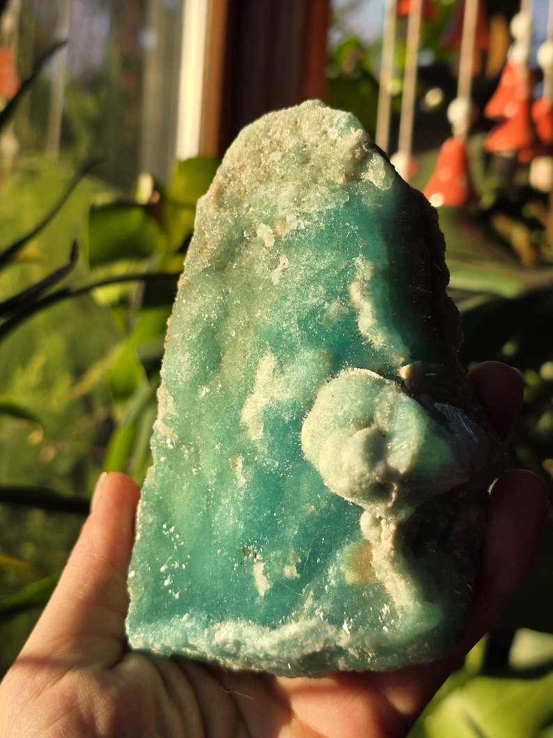 Puede incluir: Una mano sostiene un esp&eacute;cimen mineral de color turquesa con bordes &aacute;speros. La piedra tiene una superficie texturizada con parches m&aacute;s claros y una formaci&oacute;n redondeada de color m&aacute;s claro en el lado derecho. El fondo est&aacute; borroso con vegetaci&oacute;n y luz solar.