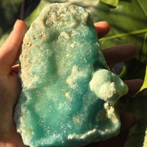 Puede incluir: Un esp&eacute;cimen mineral de color turquesa, con bordes &aacute;speros e irregulares, sostenido en una mano. La piedra tiene parches de color blanquecino. Una formaci&oacute;n m&aacute;s peque&ntilde;a y redondeada est&aacute; unida a la pieza principal.