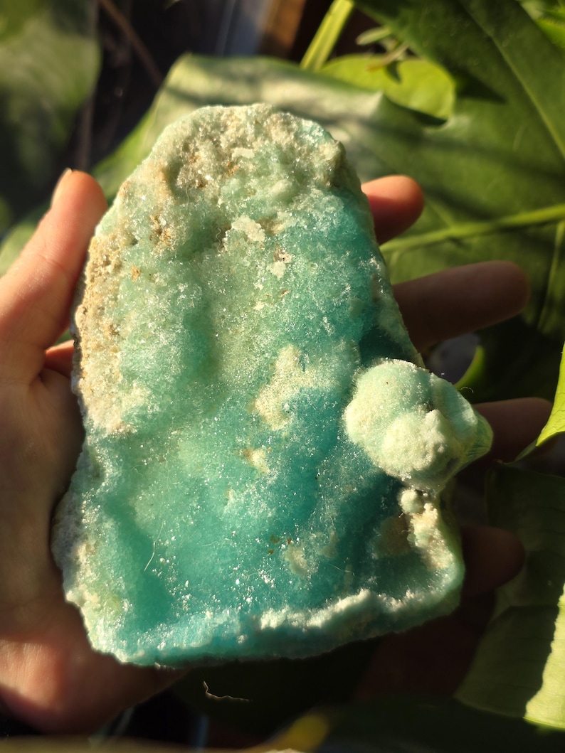 Puede incluir: Un esp&eacute;cimen mineral de color turquesa, con bordes &aacute;speros e irregulares, sostenido en una mano. La piedra tiene parches de color blanquecino. Una formaci&oacute;n m&aacute;s peque&ntilde;a y redondeada est&aacute; unida a la pieza principal.