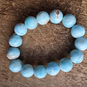 May include: A light blue beaded bracelet with round, polished beads. The bracelet is made of a series of spherical beads, each with a unique pattern of light and dark blue hues. The bracelet is displayed on a wooden surface.
