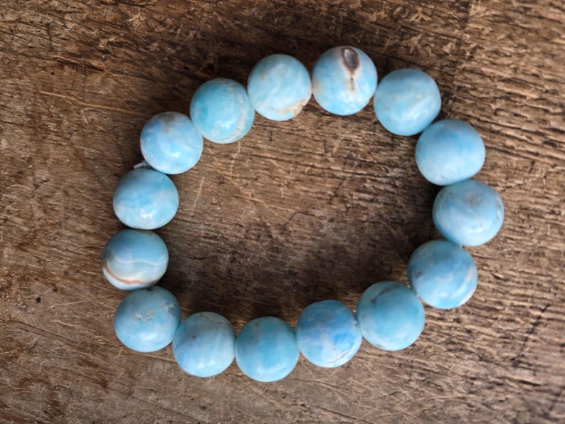 May include: A light blue beaded bracelet with round, polished beads. The bracelet is made of a series of spherical beads, each with a unique pattern of light and dark blue hues. The bracelet is displayed on a wooden surface.