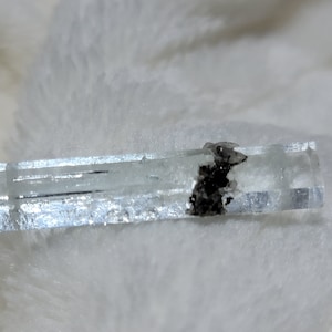May include: A light blue aquamarine crystal with a dark gray inclusion. The crystal is rectangular in shape and has a smooth, polished surface.