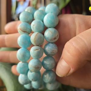 May include: A stack of three bracelets made of light blue, round beads. The beads have a marbled appearance with white and brown streaks. The bracelets are held in a hand, with a green leaf in the background.