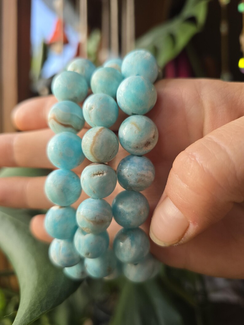 May include: A stack of three bracelets made of light blue, round beads. The beads have a marbled appearance with white and brown streaks. The bracelets are held in a hand, with a green leaf in the background.