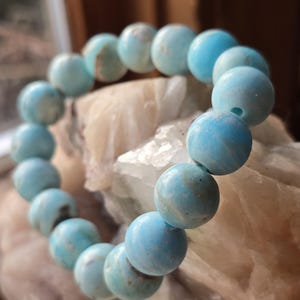 May include: A beaded bracelet featuring round, light blue beads. The beads have a slightly mottled appearance with variations in color. The bracelet is strung together, creating a circular shape. The background is a light-colored, textured surface.